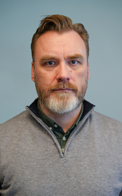 En middelaldrende mann med lys hud, kort brunt hår og heldekkende skjegg med grå striper står mot en enkel blå bakgrunn. Han har på seg en grå genser med kvart glidelås over en skjorte med krage og ser direkte inn i kameraet med et nøytralt uttrykk.