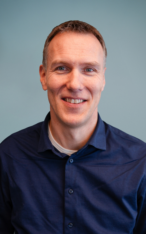 En mann med kort brunt hår smiler til kameraet og viser en følelse av føyelighet. Han har på seg en marineblå skjorte med knapper over en hvit t-skjorte med rund hals. Den enkle lyseblå bakgrunnen og det jevne lyset fremhever ansiktet hans tydelig.