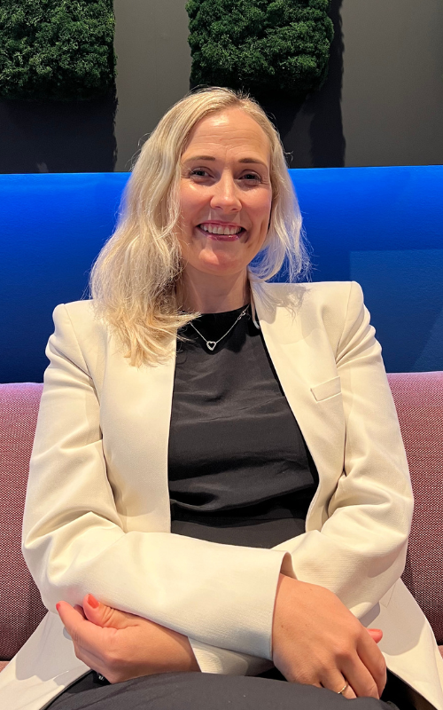 En smilende kvinne med langt blondt hår sitter i en rosa sofa på en moderne arbeidsplass. Hun har på seg en kremfarget blazer over en svart topp og folder hendene i fanget. Bak henne er det en blåbelyst vegg med grønt løvverk mot en mørk bakgrunn.