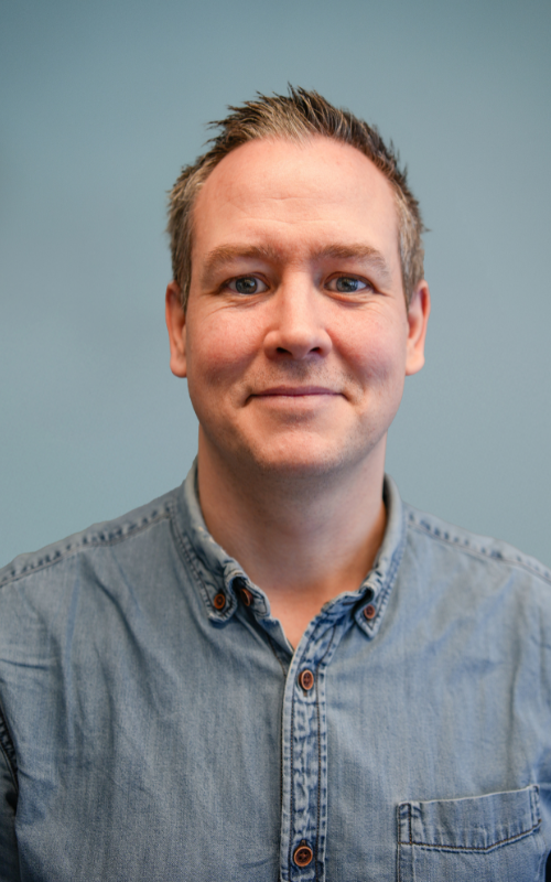 En mann med kort, lysebrunt hår og blå øyne smiler forsiktig til kameraet. Han har på seg en lyseblå denimskjorte med knapper. Bakgrunnen er ensfarget og lyseblå, noe som gir bildet et rent og profesjonelt utseende.