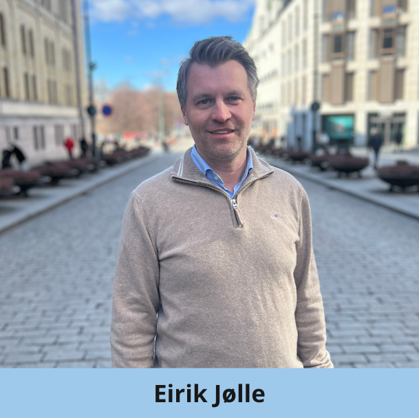 En mann med kort, brunt hår og trimmet skjegg står på en brosteinsbelagt bygate. Han har på seg en lys beige genser med glidelås over en blå skjorte med krage og smiler lett. Høye bygninger og fotgjengere står langs gaten. Nederst står det "Eirik Jølle".