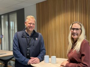 En mann og en kvinne står ved et lite rundt bord med to hvite krus og driver med kompetansebygging. Mannen er iført marineblå vest og nøkkelbånd, mens kvinnen, iført briller og rødbrun genser, står foran en lamellvegg av tre i et lyst kontormiljø.